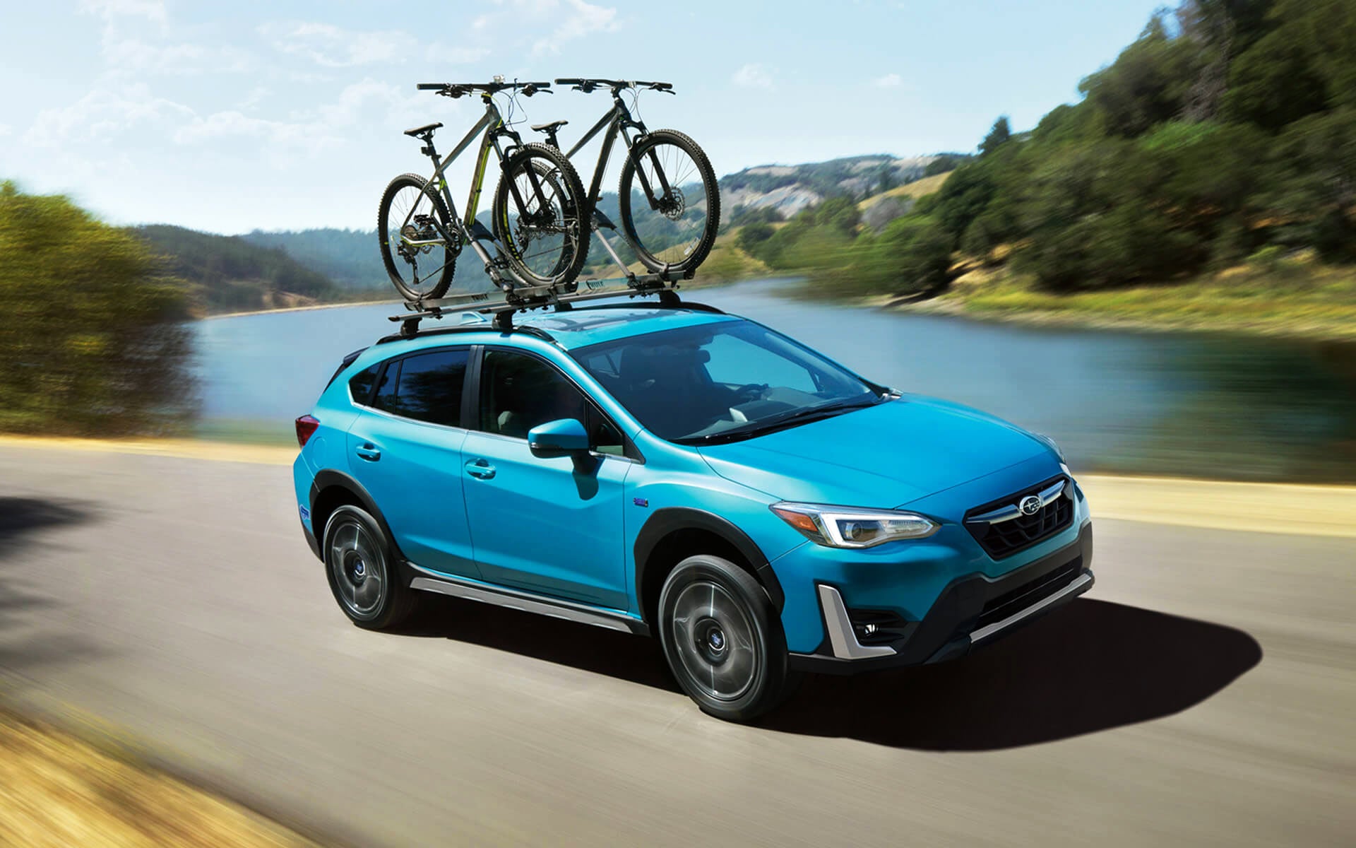 A blue Crosstrek Hybrid with two bicycles on its roof rack driving beside a river | DELLA Subaru of Plattsburgh in Plattsburgh NY